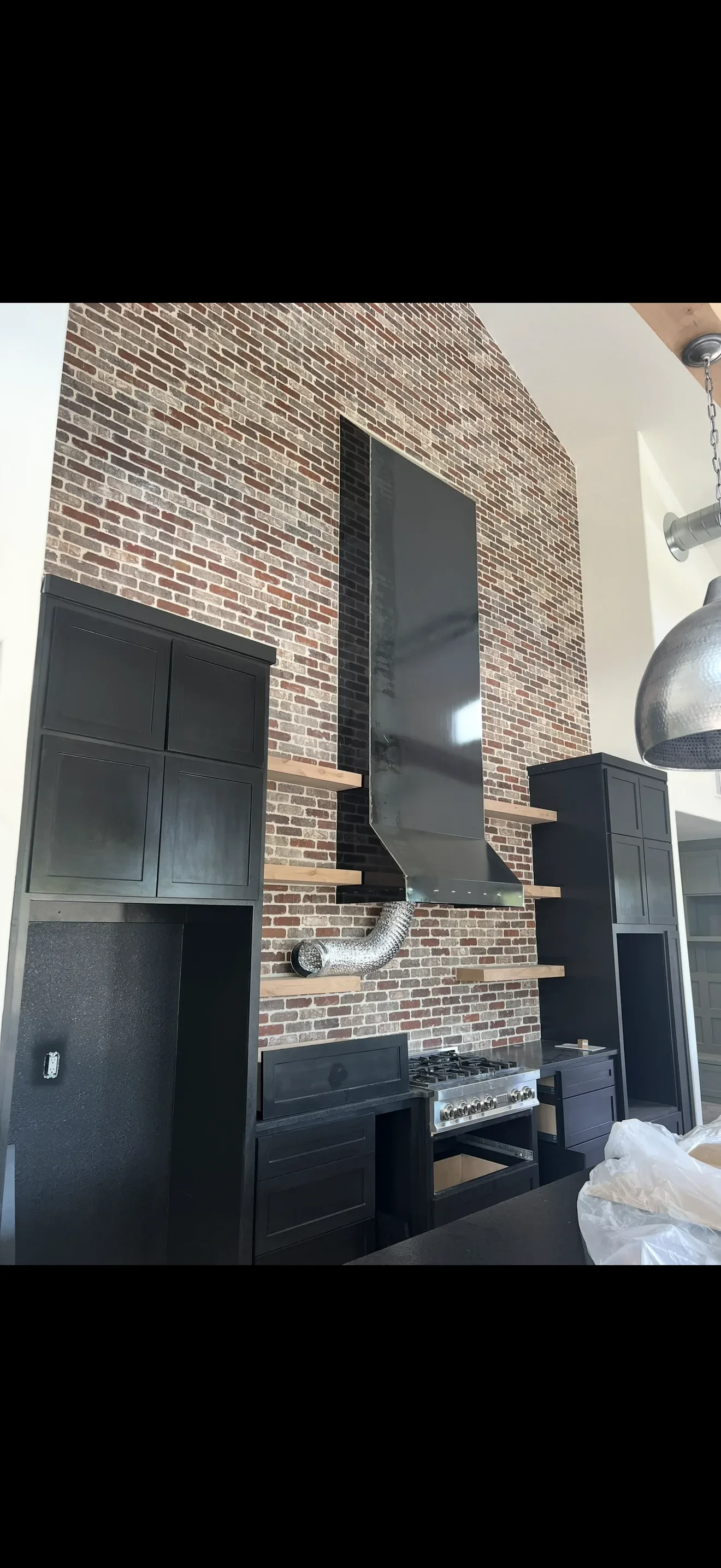 Black cabinetry and gas range against exposed red brick wall with steel hood and wooden shelves