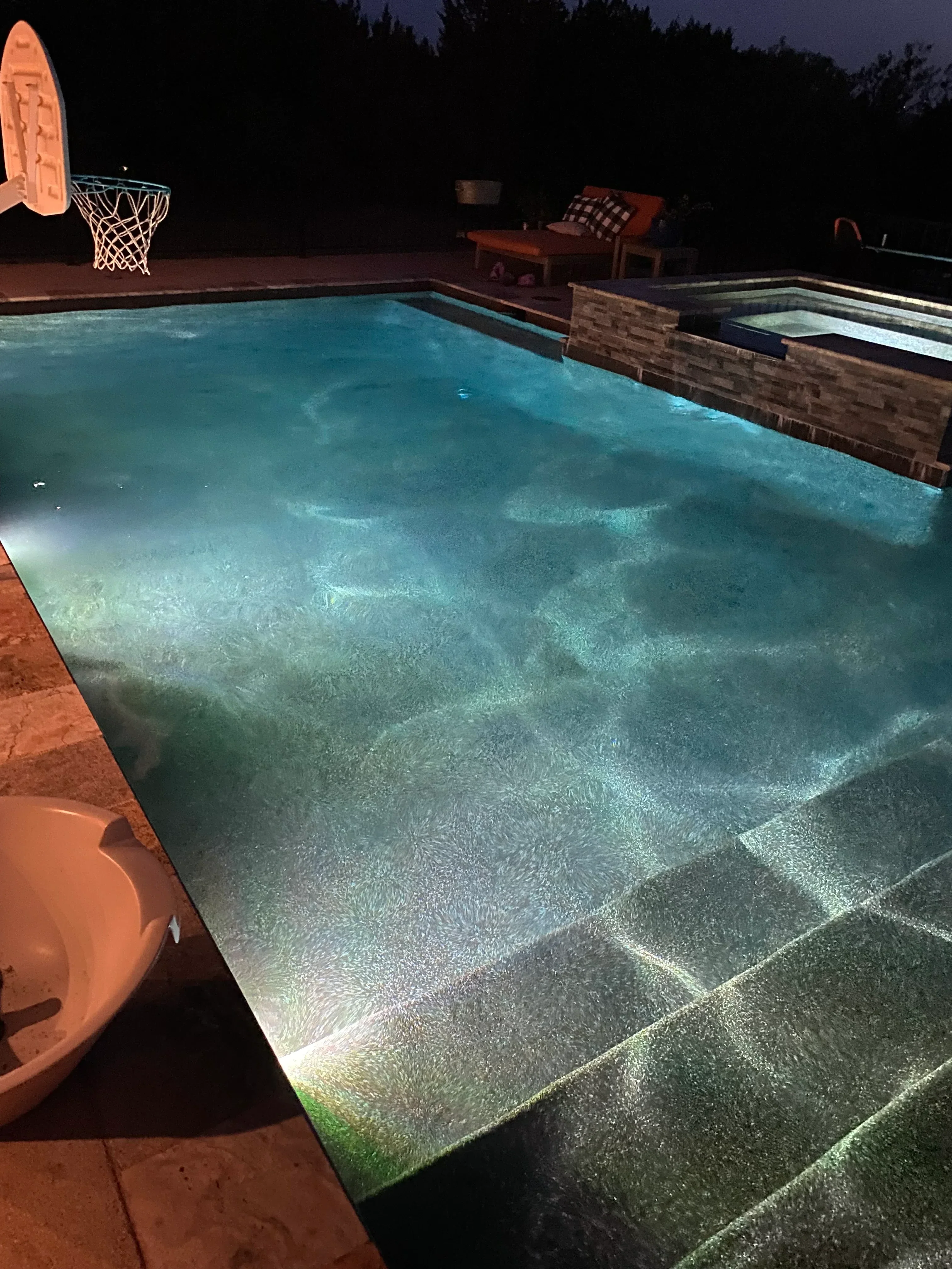 Lit pool deck with textured concrete paving, brick border, and landscape lighting at night